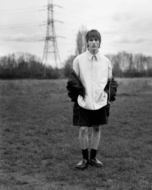 Monochrome full shot of a young adult man in a shirt and shorts standing in a field with a utility pole and cloudy sky.