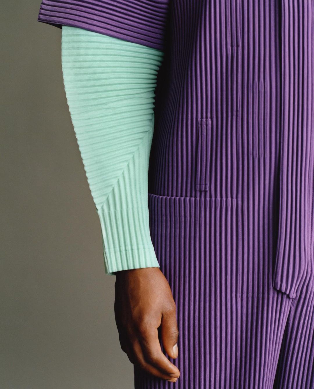 Close-up of a dark-skinned arm and hand adorned in mint-green pleated sleeve and purple pleated jumpsuit against a neutral backdrop.