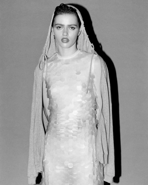 Monochrome portrait of a young adult woman in a sequin dress and hoodie, posing indoors against a plain grey background in studio lighting.