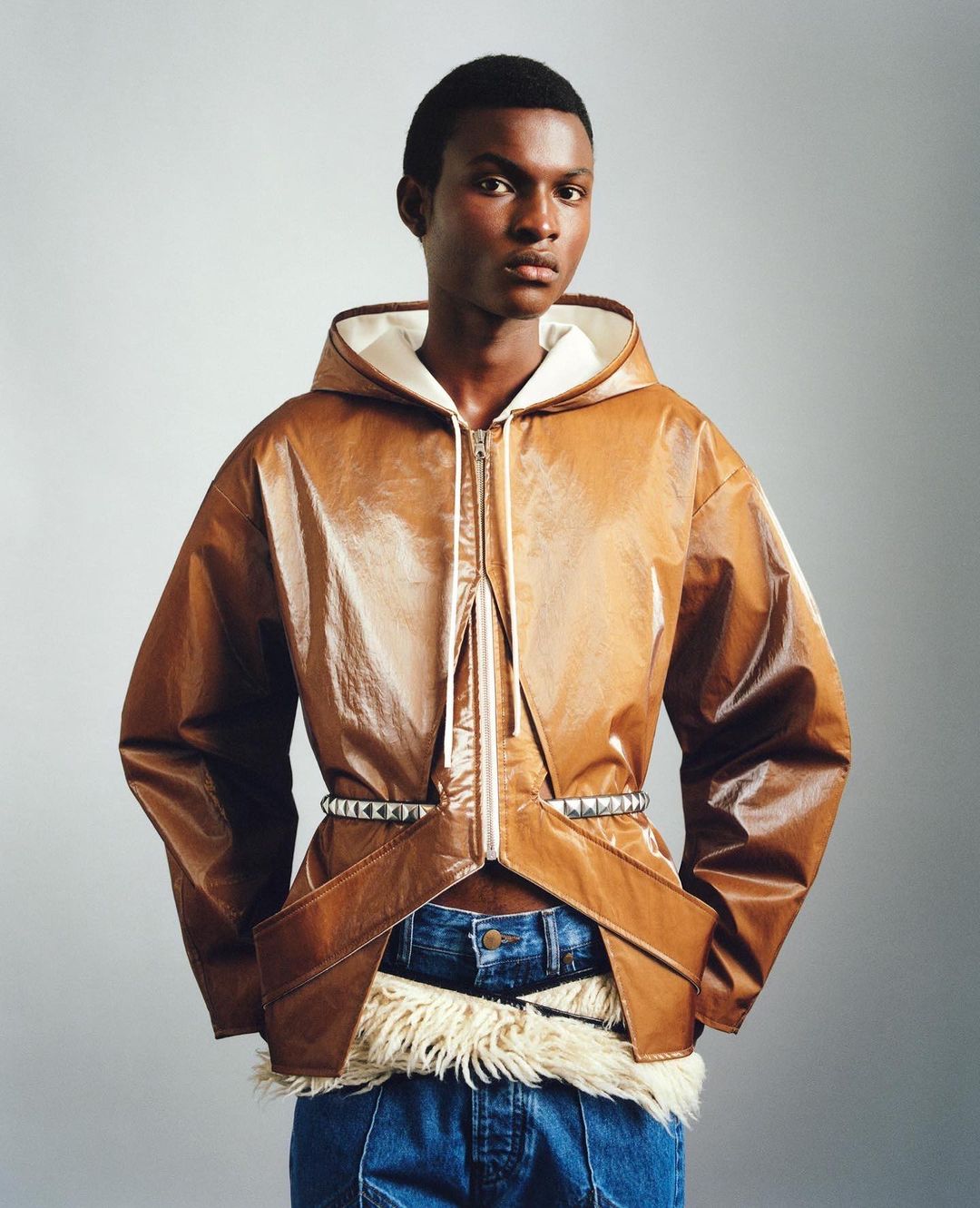 Man in a brown faux leather hooded jacket with silver studded belt and jeans, posing in front of a grey gradient backdrop.