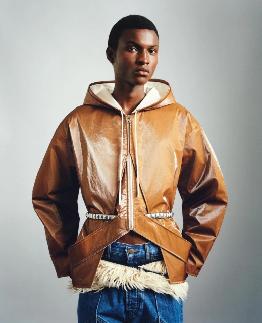 Man in a brown faux leather hooded jacket with silver studded belt and jeans, posing in front of a grey gradient backdrop.
