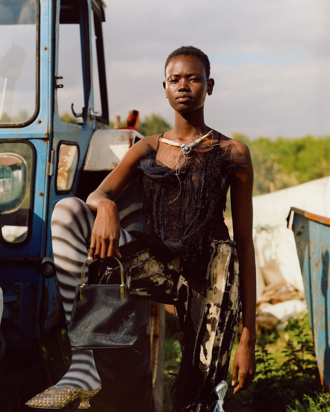 Fashion shot of a Black woman posing in front of a blue tractor, wearing a black tulle top and patterned skirt, accessorized with a necklace and purse.