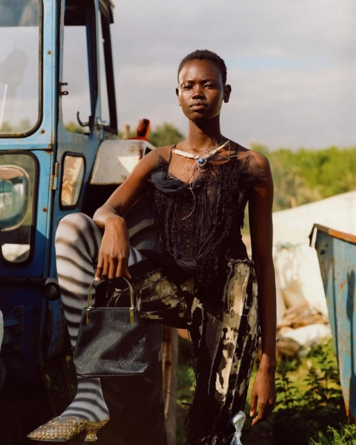 Fashion shot of a Black woman posing in front of a blue tractor, wearing a black tulle top and patterned skirt, accessorized with a necklace and purse.