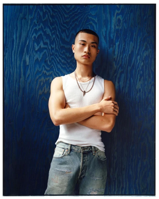 Portrait of a young adult male with short hair and crossed arms wearing a white tank top and jeans against a textured blue background.