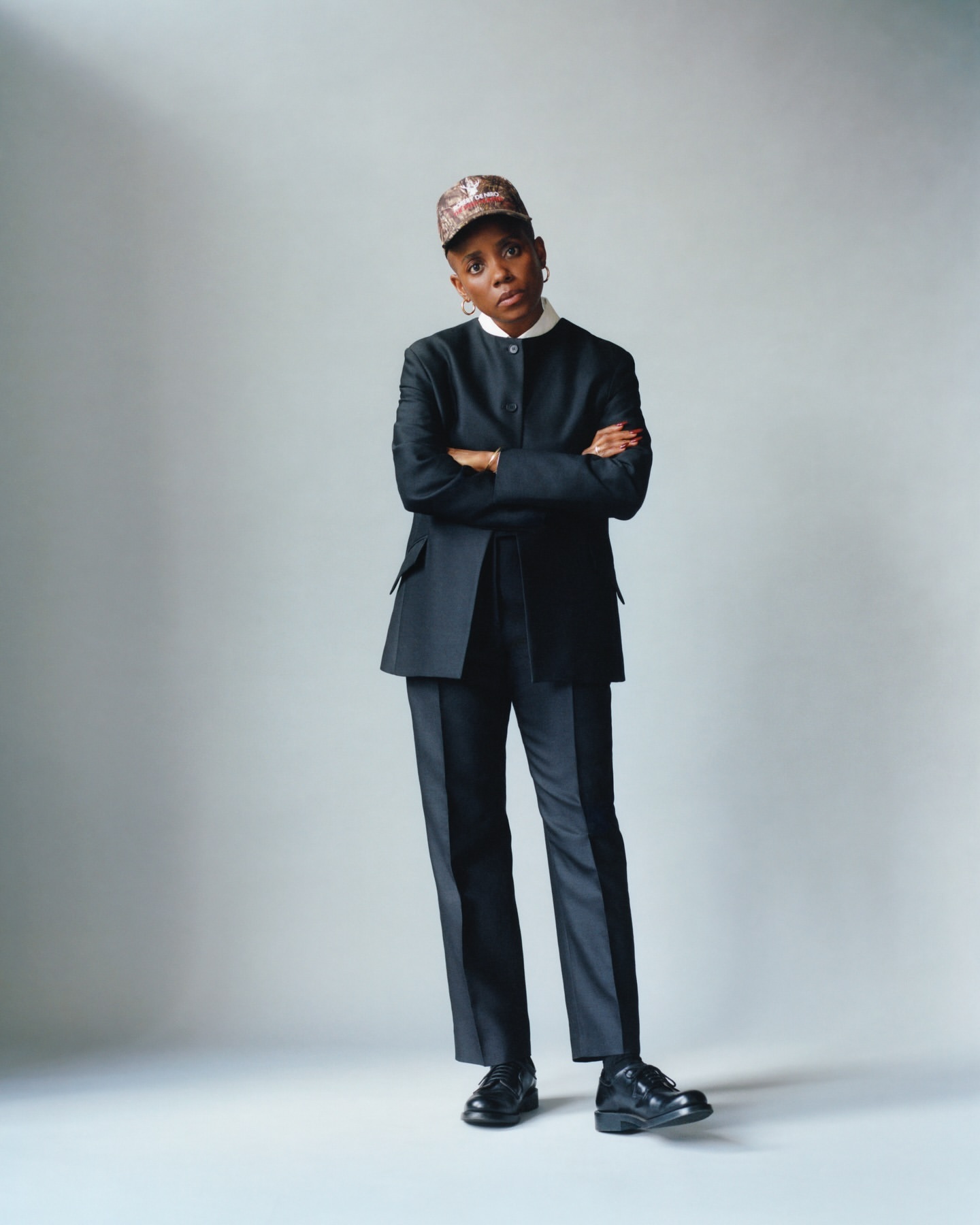 Adult woman in a black suit and patterned cap, posing with arms crossed against a neutral backdrop, showcasing professional style.