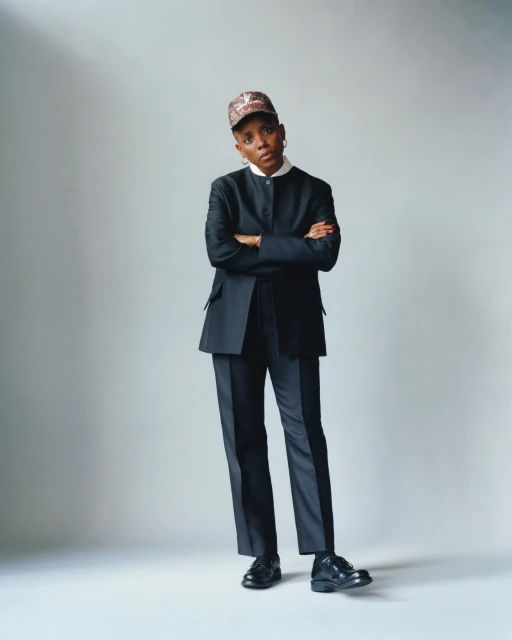 Adult woman in a black suit and patterned cap, posing with arms crossed against a neutral backdrop, showcasing professional style.