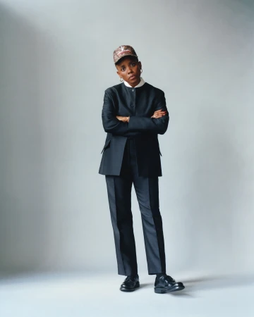 Adult woman in a black suit and patterned cap, posing with arms crossed against a neutral backdrop, showcasing professional style.