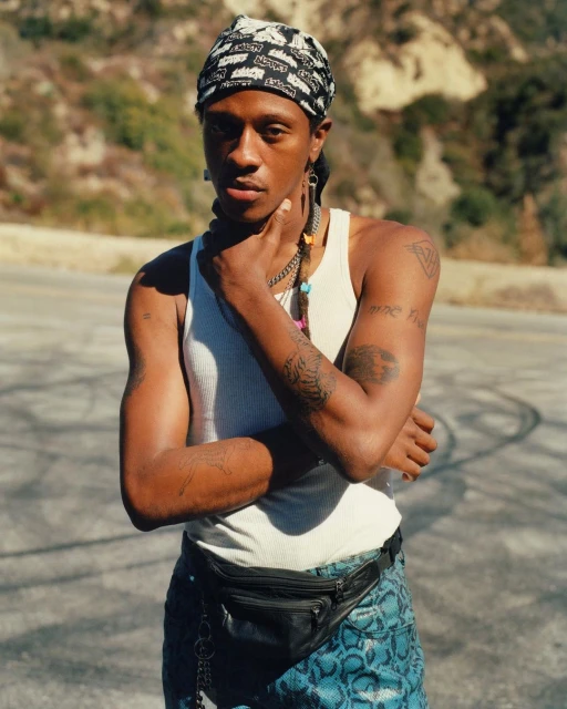 Young Black man with tattoos wears tank top, bandana, and fanny pack, posing outdoors on a road with a hillside in the background.