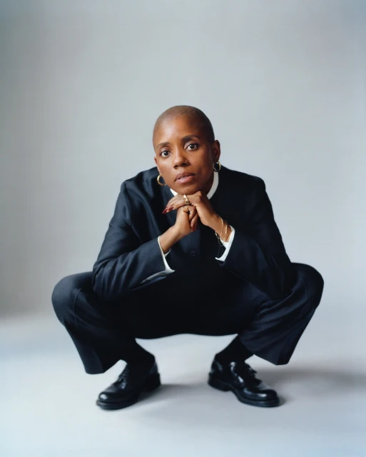 Stylish Black woman in black suit crouching with hands together, hoop earrings, short hair, neutral expression, in studio setting.