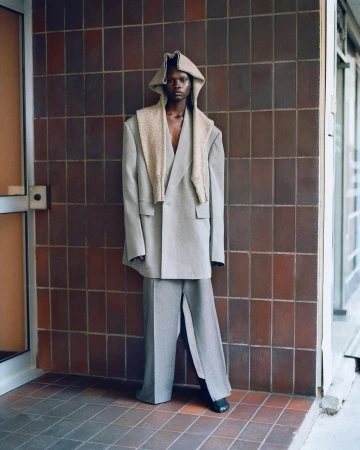 Fashionable adult in oversized gray suit stands against tiled wall, accessorized with a hood and sweater draped over the shoulders.