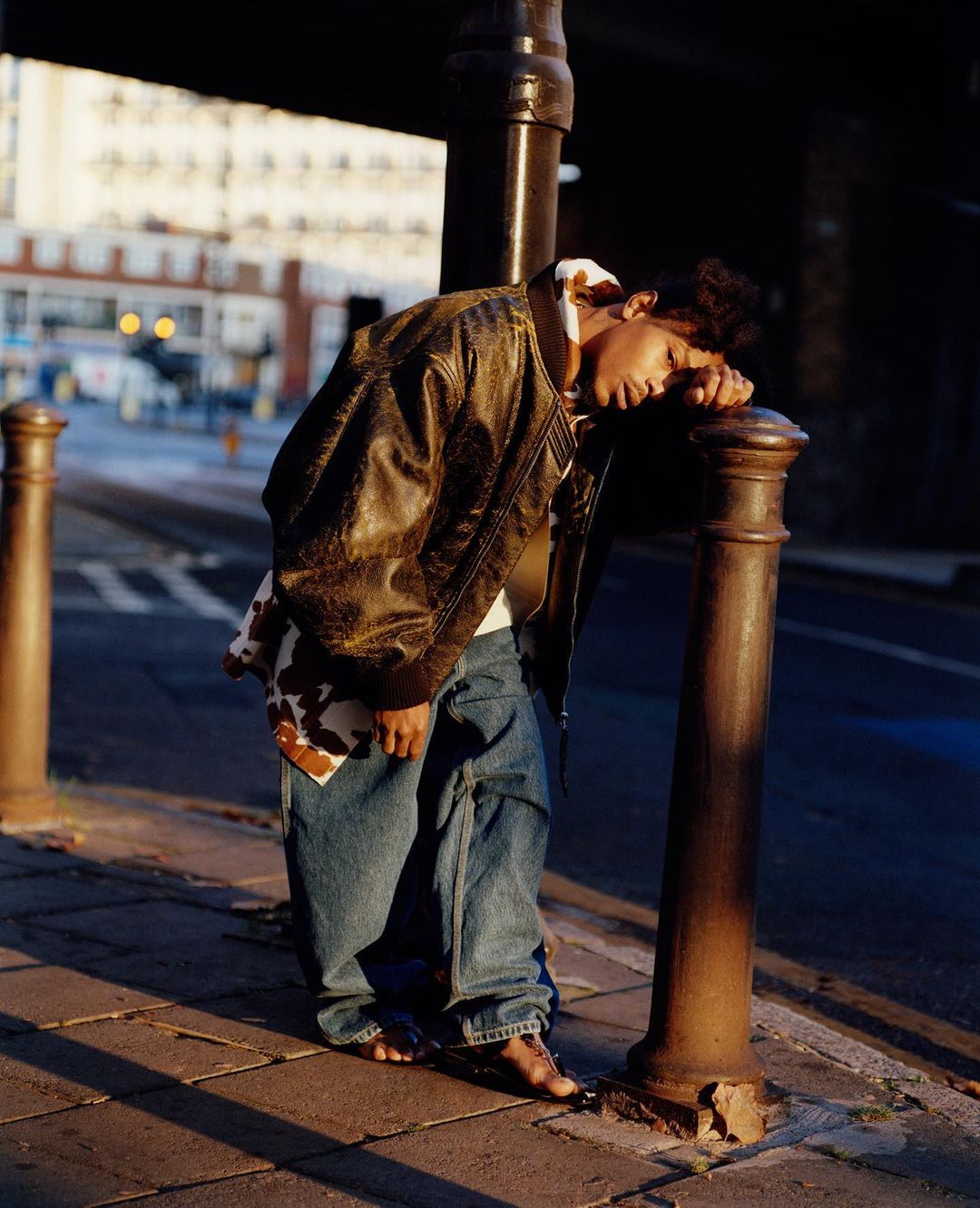 Person leaning on metal post in urban setting wearing a brown leather jacket and jeans with blurred background of city street and buildings.
