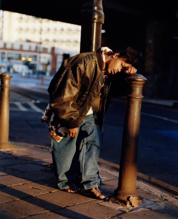 Person leaning on metal post in urban setting wearing a brown leather jacket and jeans with blurred background of city street and buildings.