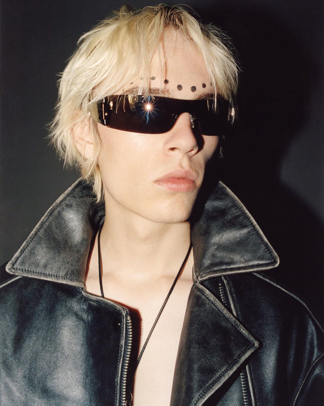 Portrait of androgynous person with sunglasses, dots on forehead, and open leather jacket in a dark photo studio setting.