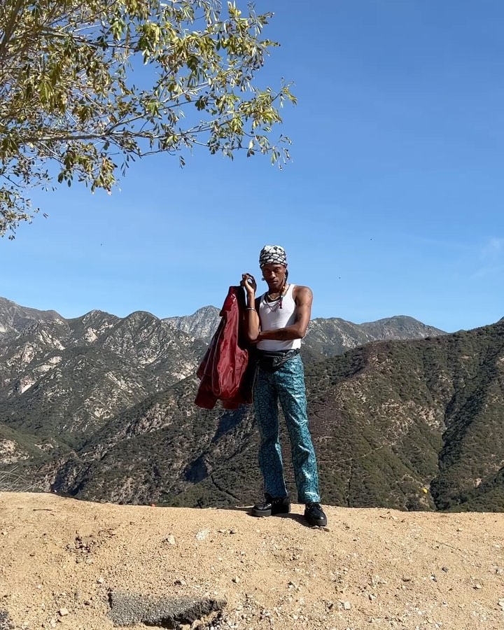 Man stands outdoors on a hill overlooking a mountain range wearing a white tank top and patterned pants, holding a red jacket.