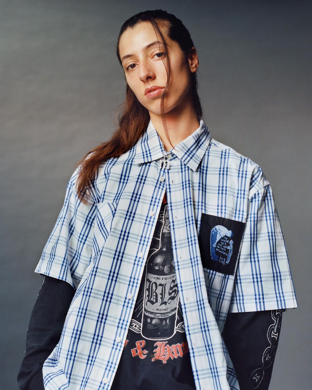 A person with long brown hair poses indoors, wearing a blue and white plaid shirt, black long sleeve shirt, and black graphic t-shirt.