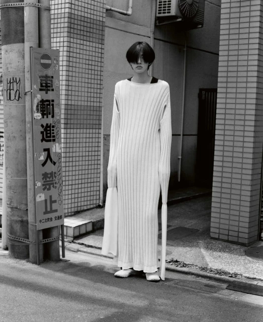 A person wearing a long white knitted dress stands on a city street in a black-and-white medium shot with a sign on a utility pole.