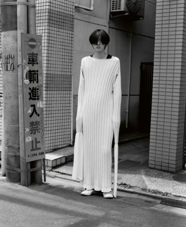 A person wearing a long white knitted dress stands on a city street in a black-and-white medium shot with a sign on a utility pole.