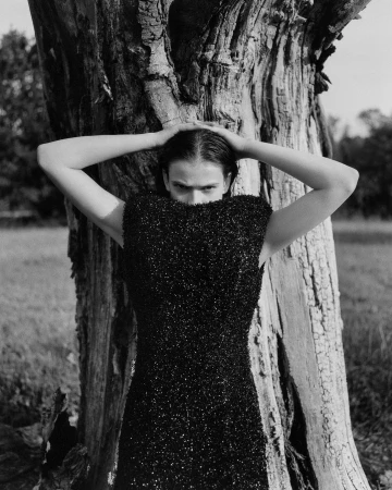 Model in black textured dress posing against a large tree trunk with hands on head, captured by Lola and Pani.