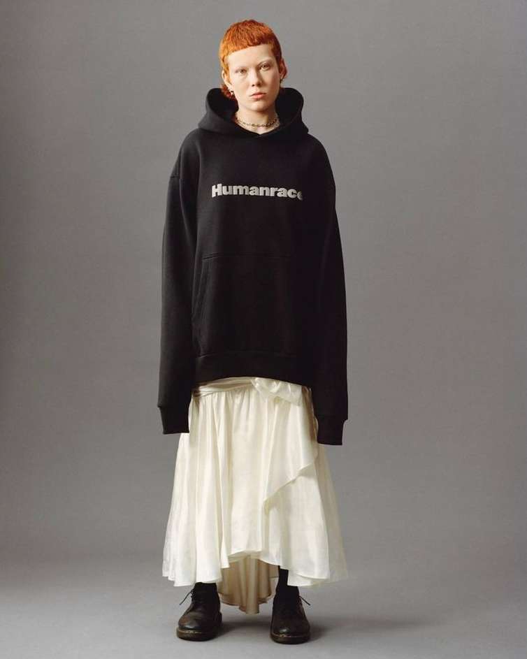 An adult female stands in a photo studio, wearing a black Humanrace hoodie, a white skirt, and black boots against a grey backdrop.