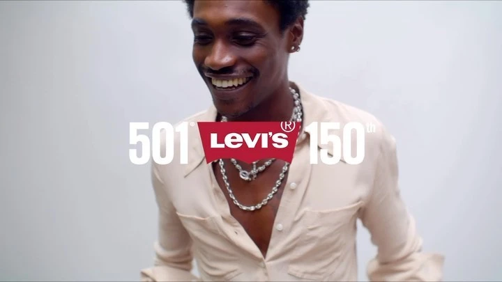 Smiling adult male poses in front of a white wall with Levi's 501 150th-anniversary branding and wears a light-colored shirt and silver necklaces.