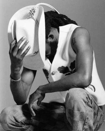 Black man wearing white hat and patterned vest kneeling against a grey backdrop