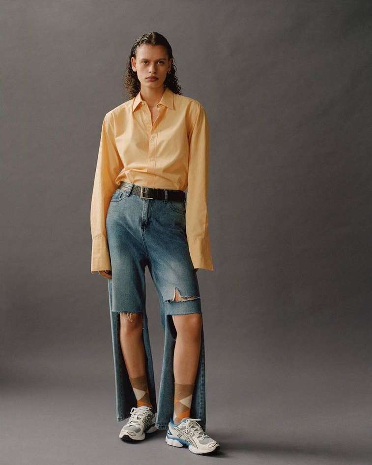 Editorial fashion shot featuring a female model in a yellow blouse and deconstructed denim pants, posing against a neutral grey background.