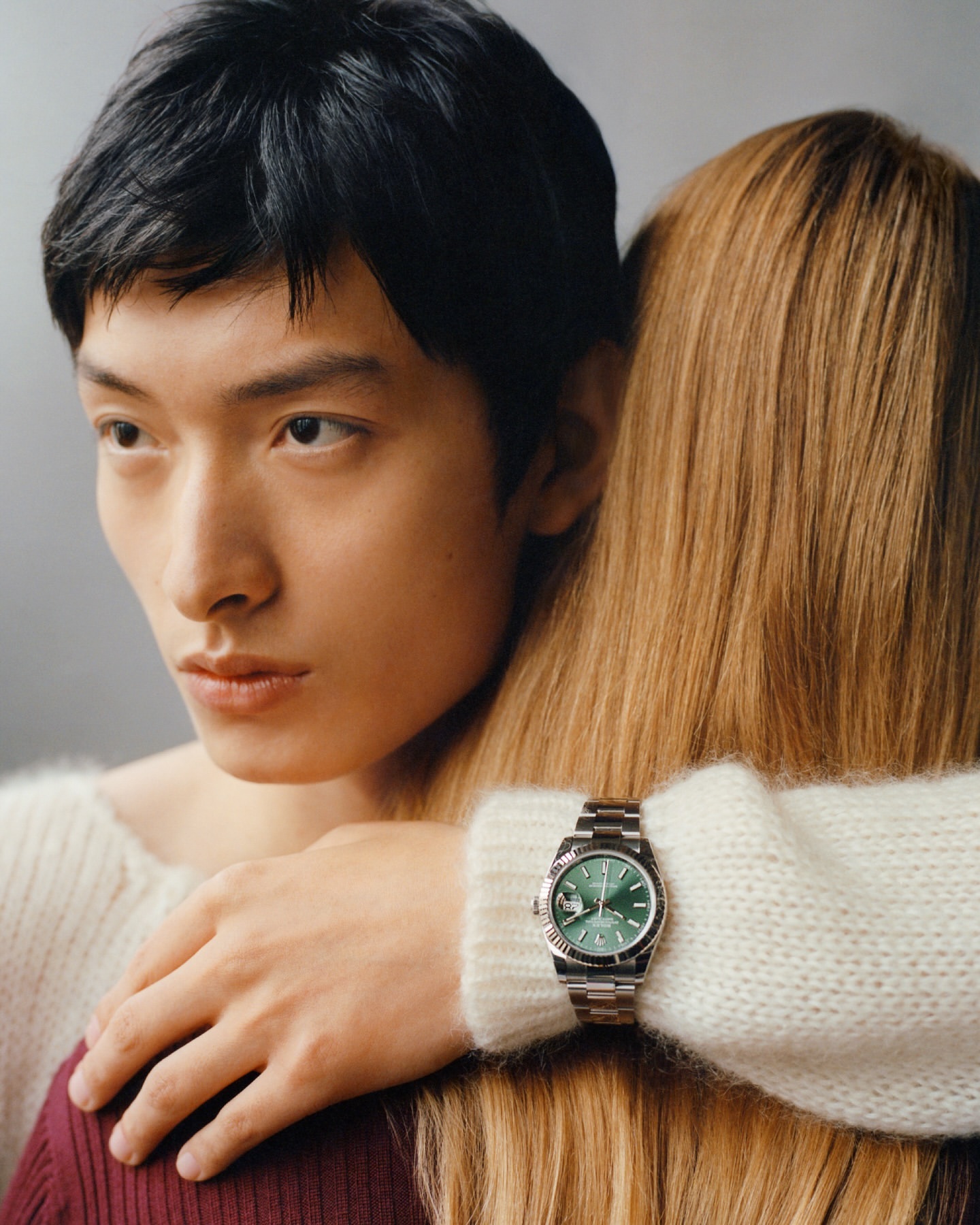 A close-up studio shot features an Asian man and a blonde woman, highlighting a green-faced wristwatch on the woman's arm.