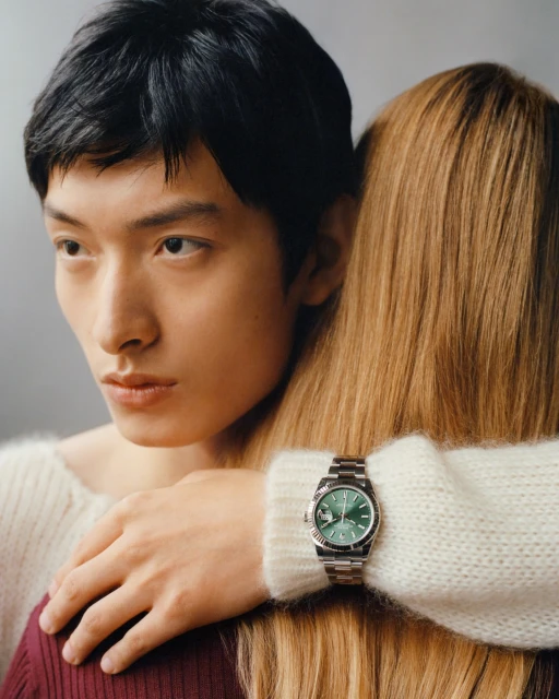 A close-up studio shot features an Asian man and a blonde woman, highlighting a green-faced wristwatch on the woman's arm.