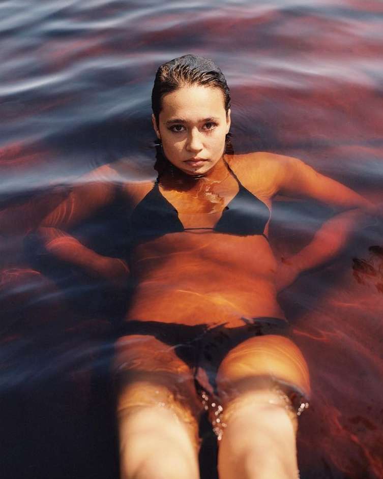 Woman in black bikini waist-deep in dark water with ripples, looking at camera, hands on hips, outdoor setting, natural light.