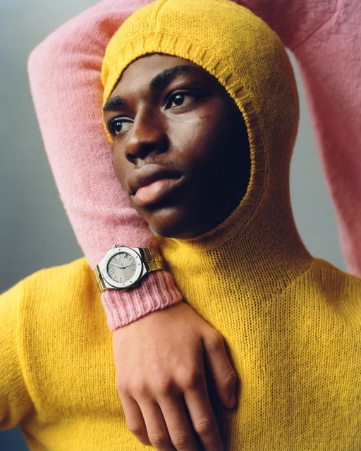 Portrait of a young Black man in a yellow turtleneck and hood, with an arm wearing a silver wristwatch resting on his shoulder.