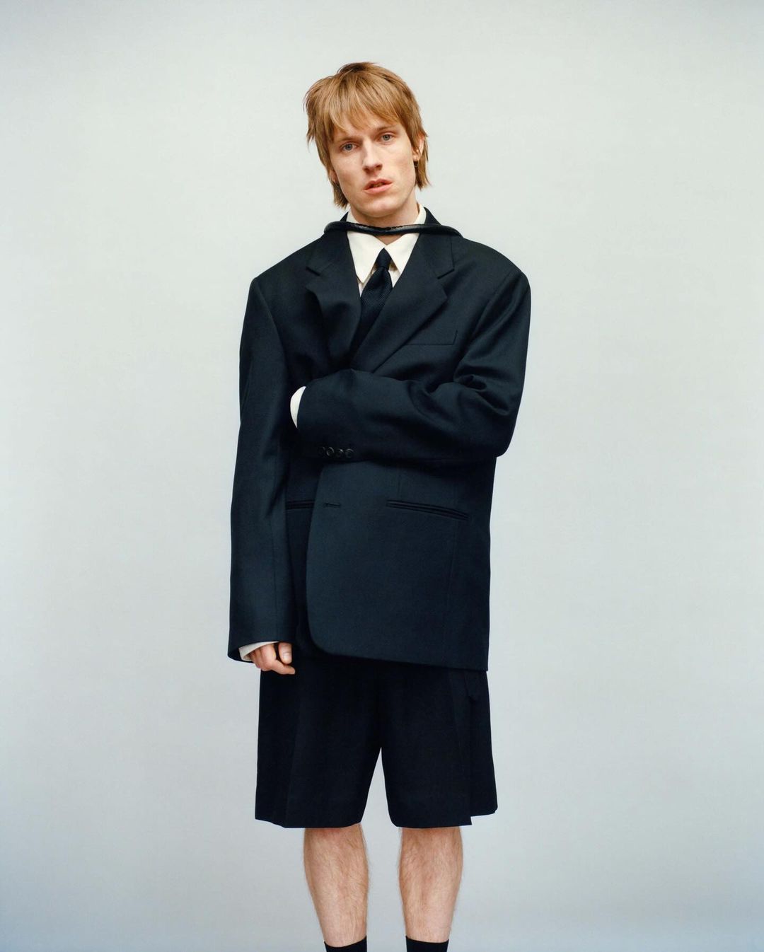 Young man with blonde hair wearing an oversized black suit jacket and shorts against a neutral studio backdrop