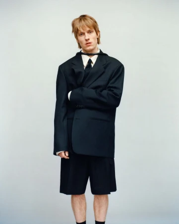 Young man with blonde hair wearing an oversized black suit jacket and shorts against a neutral studio backdrop