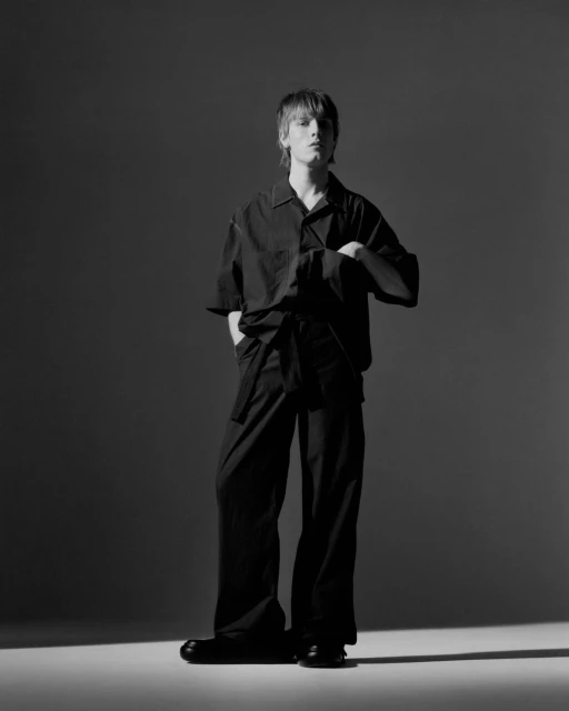 Studio shot of a male adult wearing a black outfit with an oversized shirt and wide-leg pants, standing against a neutral background.
