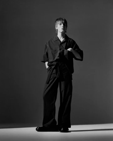 Studio shot of a male adult wearing a black outfit with an oversized shirt and wide-leg pants, standing against a neutral background.