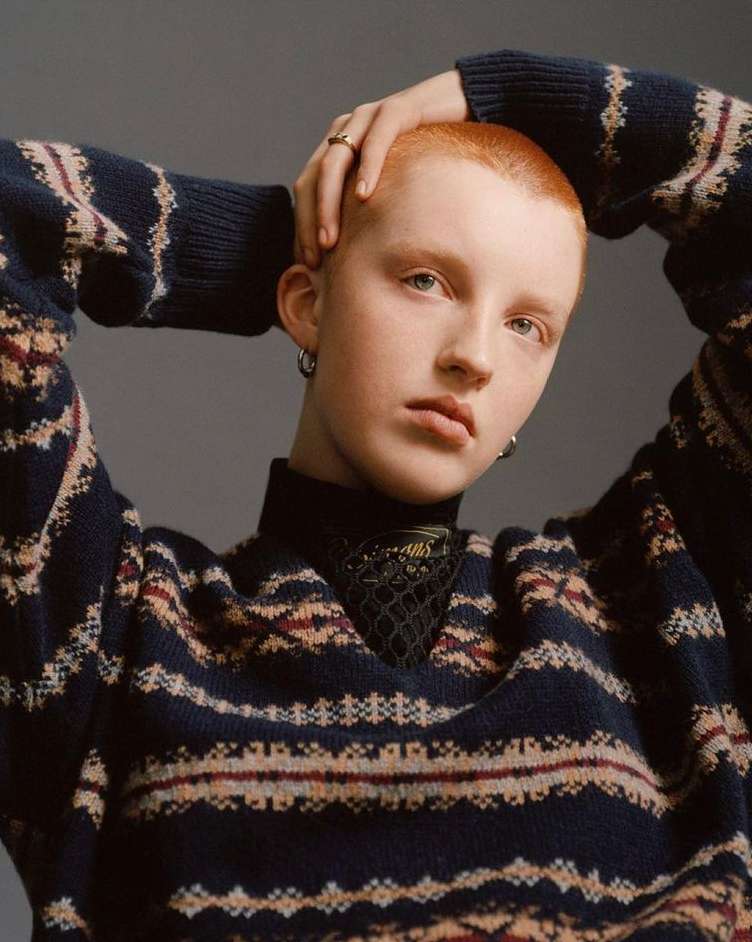 Close-up studio portrait of a short, copper-haired adult with light skin wearing a patterned sweater and jewelry, against a grey background.