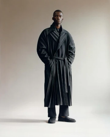 Black man wearing a long pleated black belted coat standing against a light grey studio background.