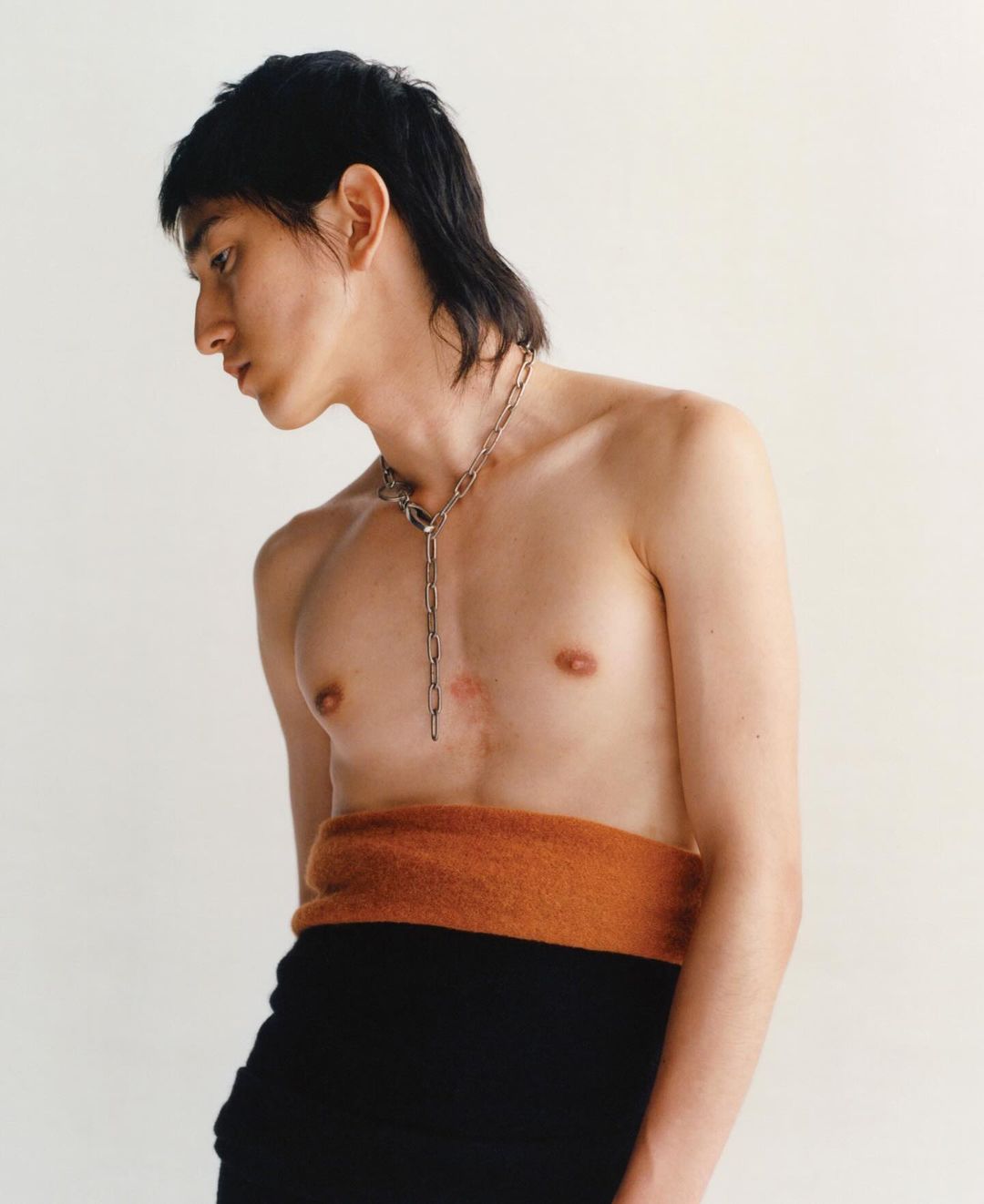 Portrait of a shirtless person with a chain posing in a studio. The person has dark hair and wears black bottoms with an orange fabric belt.