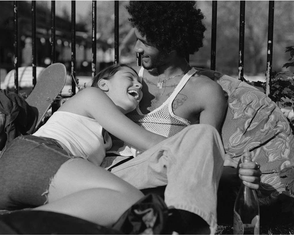 Monochrome shot of a young adult couple sitting outdoors, laughing and embracing near a metal fence with a skateboard.