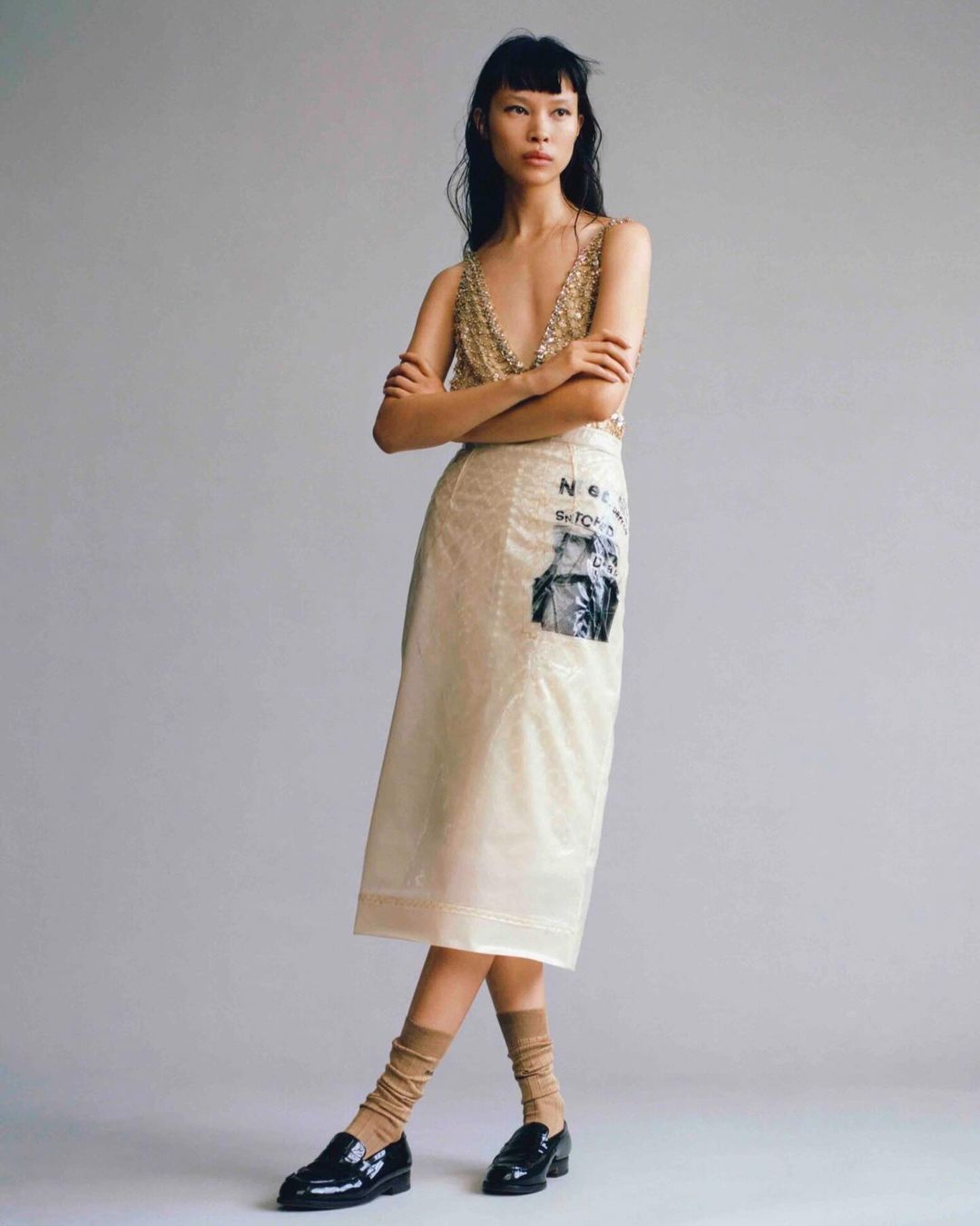 Asian fashion model in a sequin top and skirt poses in front of a plain studio backdrop with dark loafers and tan socks in a studio shot.