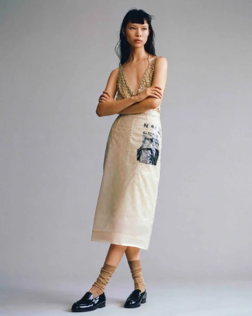 Asian fashion model in a sequin top and skirt poses in front of a plain studio backdrop with dark loafers and tan socks in a studio shot.