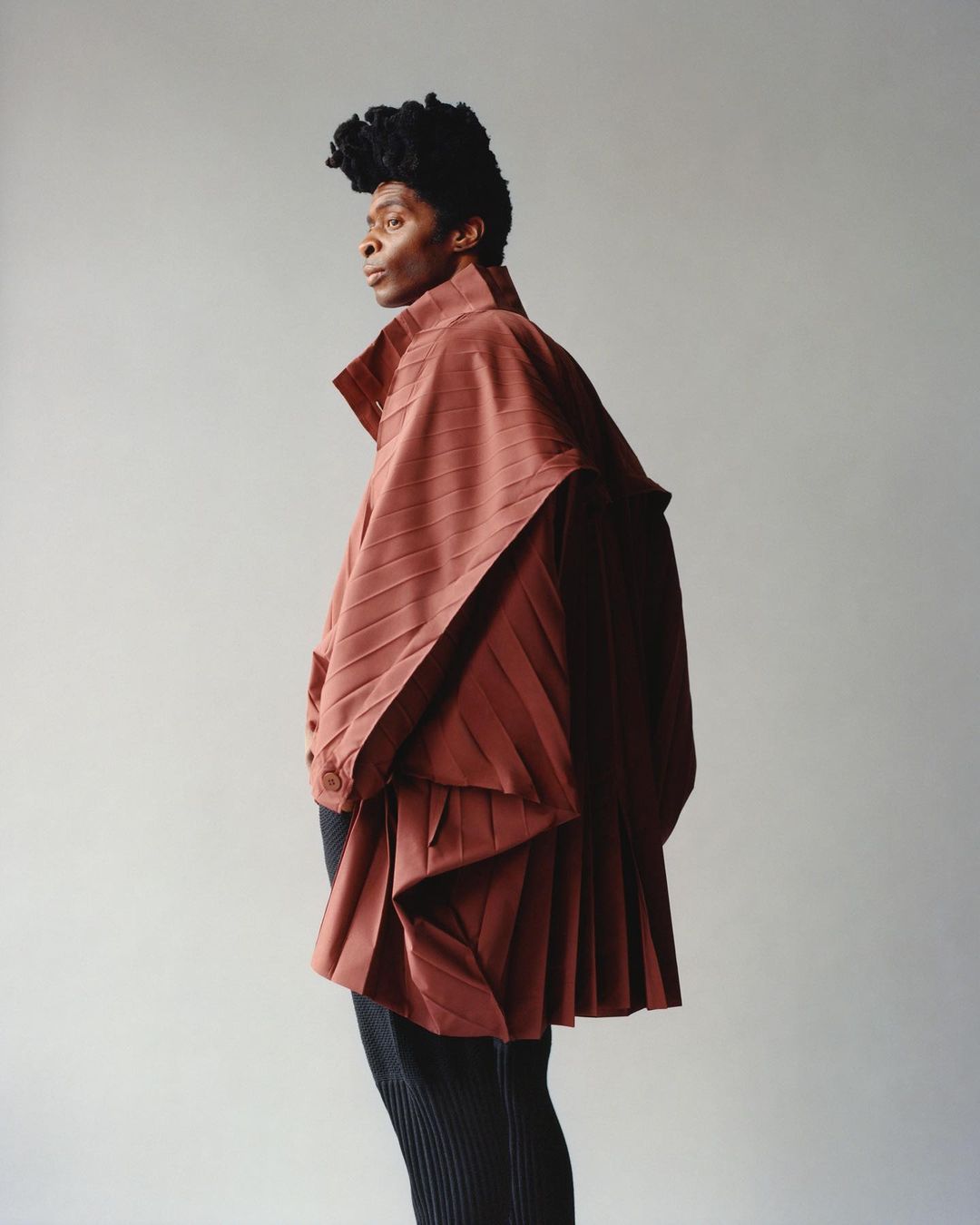 Fashion portrait of a person in a draped, pleated reddish-brown garment and black pants posing against a plain light gray background.