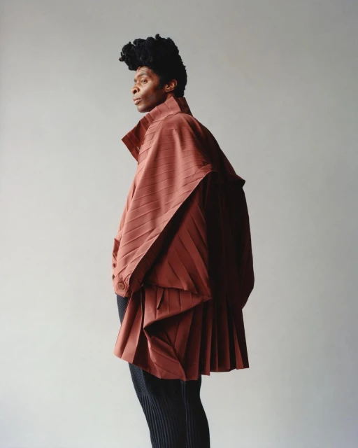 Fashion portrait of a person in a draped, pleated reddish-brown garment and black pants posing against a plain light gray background.