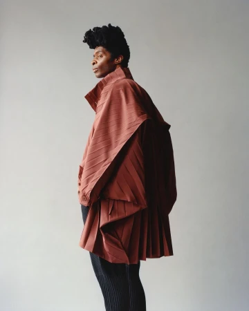 Fashion portrait of a person in a draped, pleated reddish-brown garment and black pants posing against a plain light gray background.
