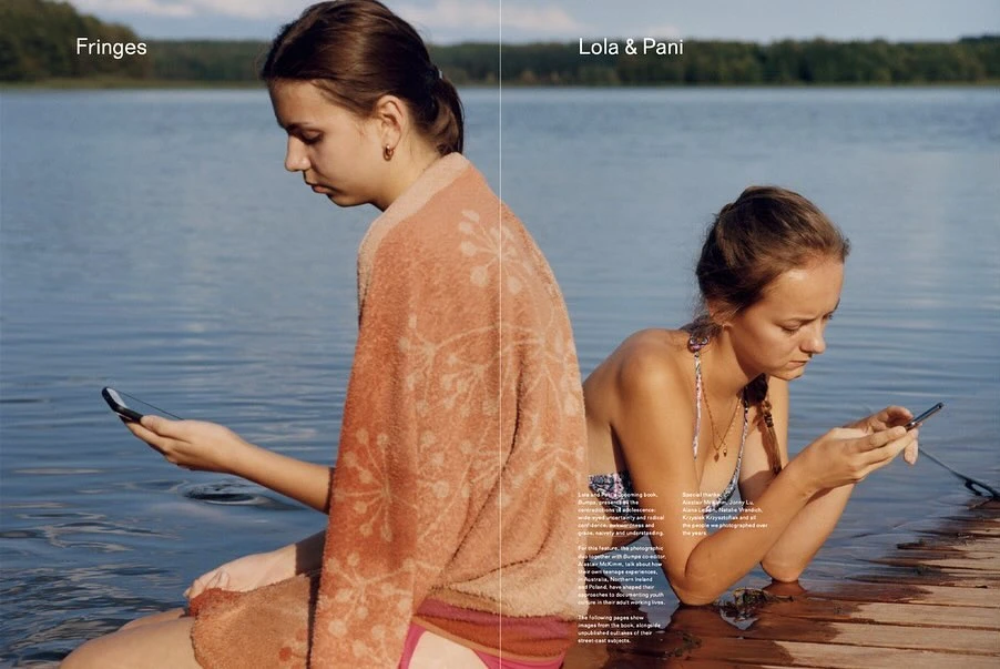 Two young women near a lake, engrossed in their mobile phones, one wrapped in an orange towel, against a backdrop of water and trees.