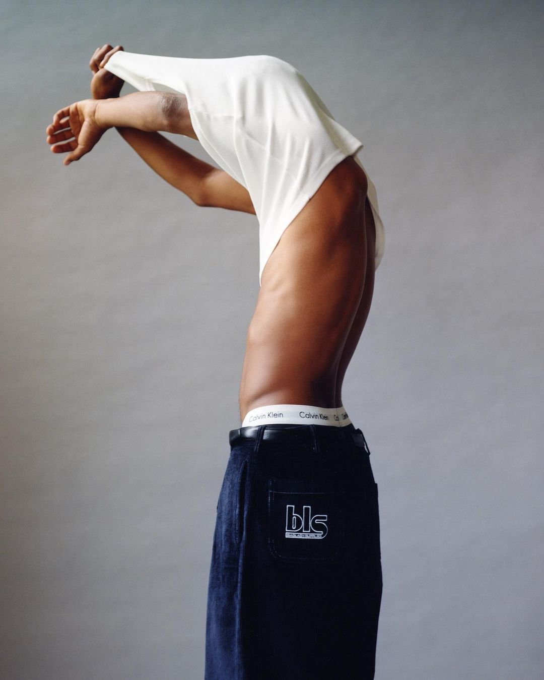 A slender, dark-skinned person in dark jeans with a Calvin Klein waistband and a white t-shirt partially removed, against a plain backdrop.