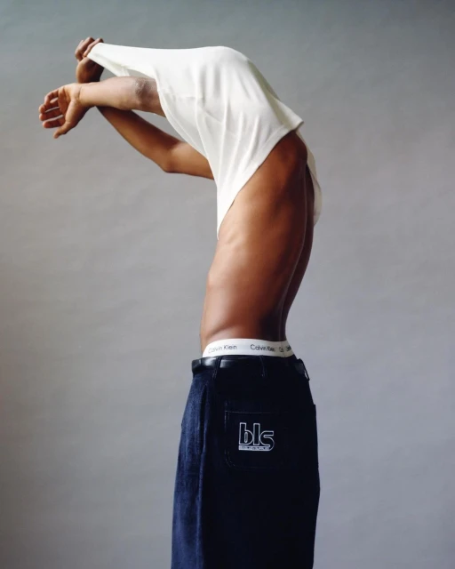 A slender, dark-skinned person in dark jeans with a Calvin Klein waistband and a white t-shirt partially removed, against a plain backdrop.