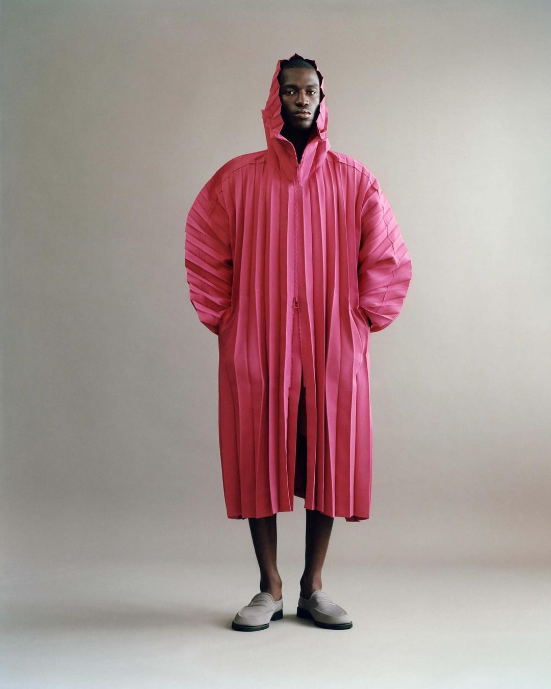Full-length studio shot of a male fashion model in a pleated, hooded, magenta overcoat with his hands in his pockets and wearing gray loafers.