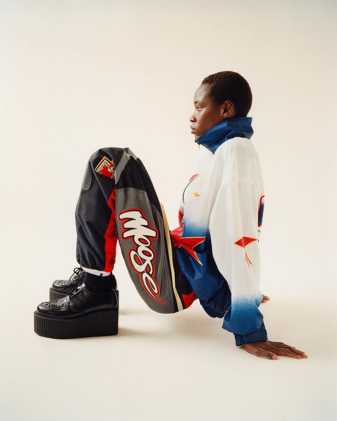 Profile shot of Black woman in racer jacket and black platform shoes against a white backdrop. Sporty fashion editorial.