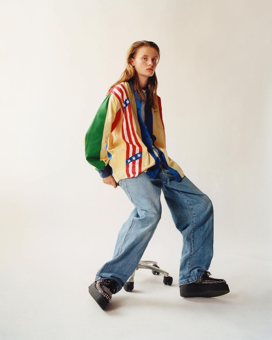Young woman in a statement jacket with a US flag motif, styled with wide-leg jeans and platform shoes, seated indoors in a fashion editorial shot.