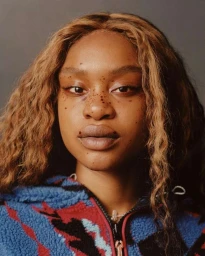 Close-up portrait of a woman with long wavy brown hair, freckles, and a blue and red patterned fleece jacket against a grey background